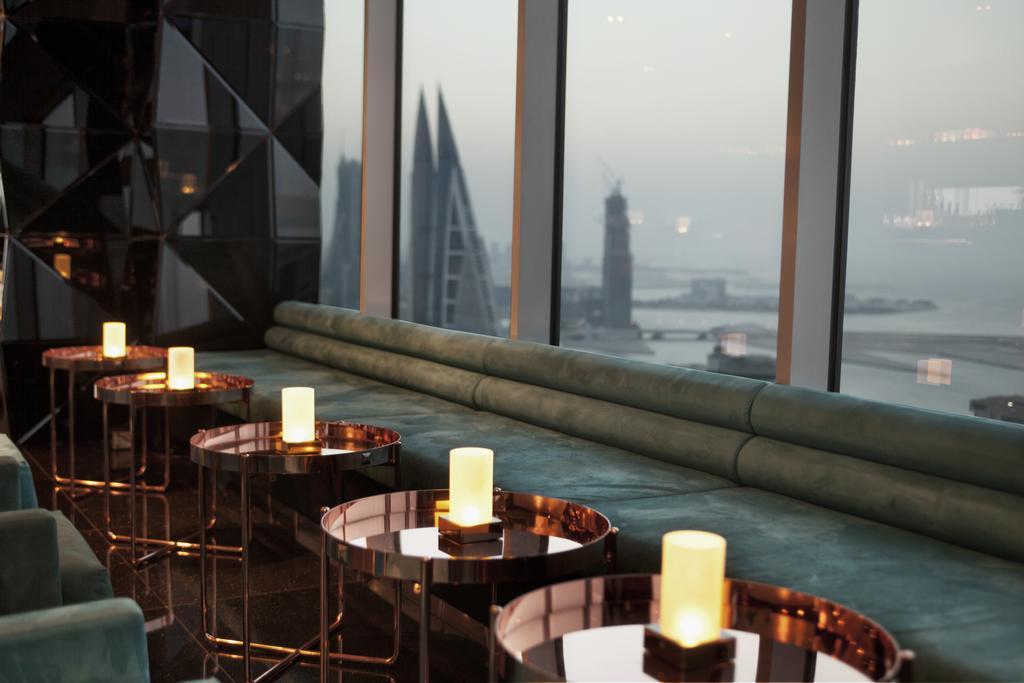 The Domain Bahrain Hotel And Spa - Adults Friendly 16 Years Plus Manáma Kültér fotó The photo depicts a stylish lounge area with a modern aesthetic. There are several circular tables made of metallic materials, each topped with a small glowing candle. A long, plush seating area in a teal color runs along the window, offering a view 